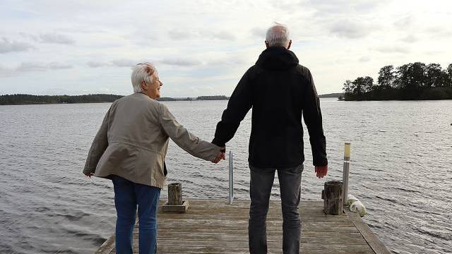 Två äldre vuxna står på en träbrygga med utsikt över en sjö och håller varandra i handen. Himlen är molnig och träd syns i fjärran.