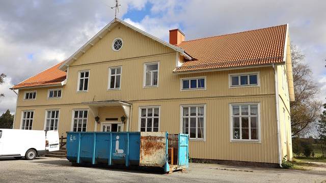 En stor gul träbyggnad med rött tegeltak, vitmålade fönster och en blå industricontainer på parkeringen utanför.
