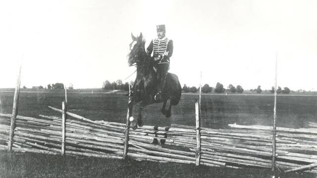 En uniformerad soldat till häst hoppar över ett trästaket på ett öppet fält.