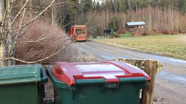 Två soptunnor står i förgrunden nära en grusväg, en lastbil kör iväg och i bakgrunden syns hus omgivna av träd.