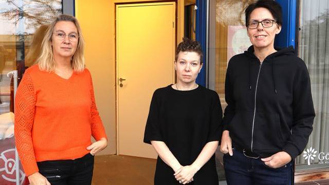 Tre personer står utanför en entré till en byggnad och poserar för fotografering. Två bär glasögon och alla är klädda i vardagskläder.