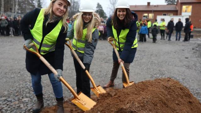 Tre kvinnor i skyddsvästar och hjälmar håller i spadar och tar första spadtaget på en byggarbetsplats, med en folkmassa och byggnader i bakgrunden.