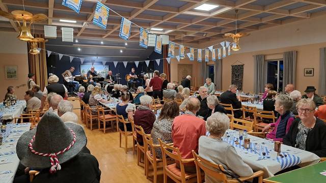 En grupp människor sitter vid långbord i en dekorerad hall och ser ett band uppträda på scenen. Blå och vita banderoller hänger från taket.