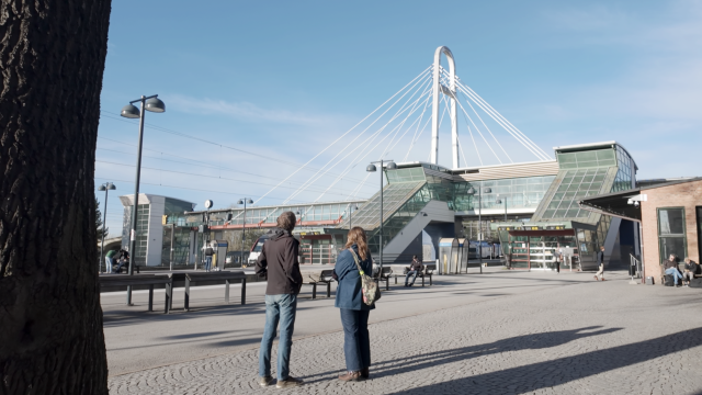 Två personer står vända mot en modern tågstation i glas med en karakteristisk snedkabelbro för fotgängare under en klarblå himmel.