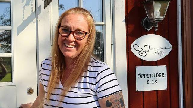 En kvinna med långt blont hår och glasögon, klädd i en randig skjorta, står leende i en dörröppning bredvid en skylt med öppettider.