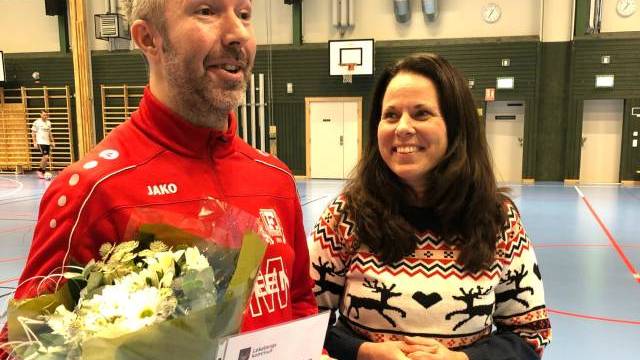 En man i röd sportjacka håller i en bukett och ett diplom och står bredvid en leende kvinna i en tröja med vintertema i en gymnastiksal.