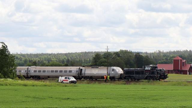 Ett tåg och två underhållsfordon står stilla på spåren i ett landsbygdsområde med gröna fält, träd och en röd lada i bakgrunden. En vit skåpbil och tre personer befinner sig i närheten.