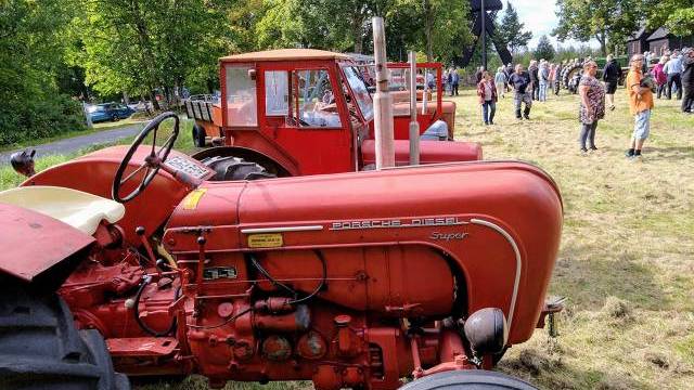 Två röda vintagetraktorer står parkerade på gräset vid ett utomhusevenemang, med människor samlade och träd i bakgrunden.