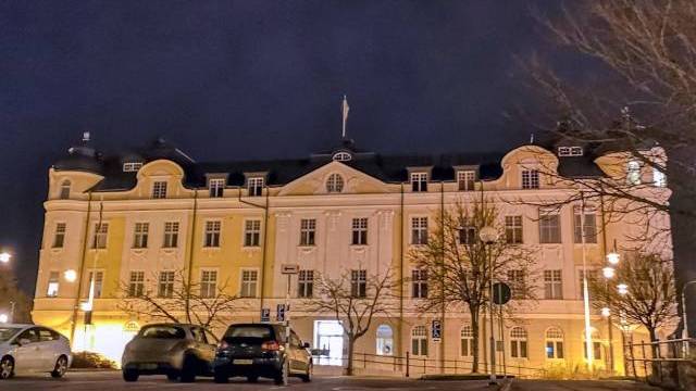 En stor, upplyst byggnad med välvda fönster och mörkt tak syns på natten, med parkerade bilar och lövlösa träd framför.