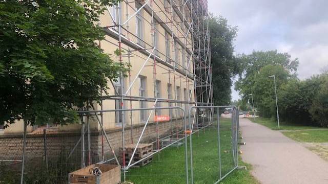 En byggnad under uppförande är omgiven av byggnadsställningar och ett tillfälligt metallstaket, med lastpallar och byggmaterial på gräset i närheten. En asfalterad stig löper längs med platsen.