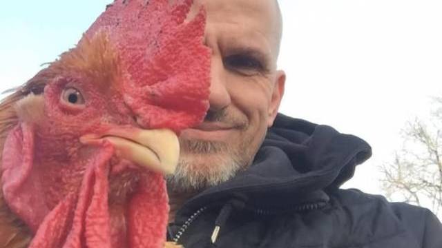 En man i svart jacka står utomhus och håller en tupp nära sitt ansikte så att deras ögon är i höjd med varandra, vilket skapar en jämförelse sida vid sida.