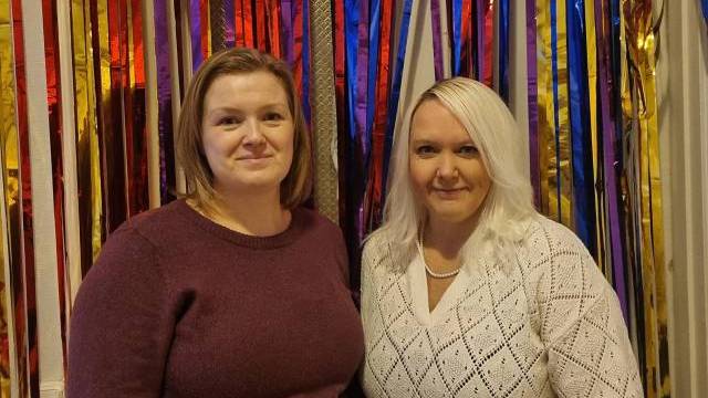 Två kvinnor står sida vid sida framför färgglada metalliska serpentiner, den ena bär en rödbrun tröja och den andra en vit tröja.