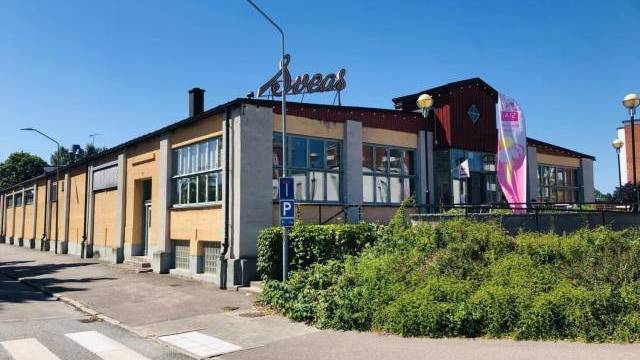 En stor byggnad i industriell stil med gula tegelväggar, stora fönster och en metallskylt på taket, belägen vid ett soligt gathörn med grönska och en flagga.