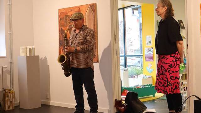 En man spelar saxofon medan en kvinna står bredvid i ett konstgalleri, med konstverk på väggarna och en öppen dörr som visar ett färgglatt rum bortom.