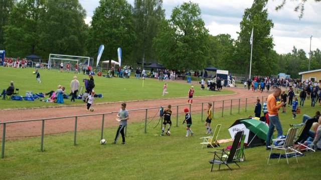 Barn och vuxna är samlade på en gräsbeklädd idrottsplats med pågående fotbollsmatcher och åskådare som sitter eller står längs sidlinjen.