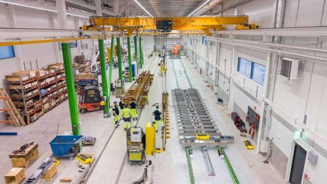 Stor industriverkstad med arbetare i varselkläder, maskiner, lagerhyllor och traverskran i en ljus och modern anläggning.