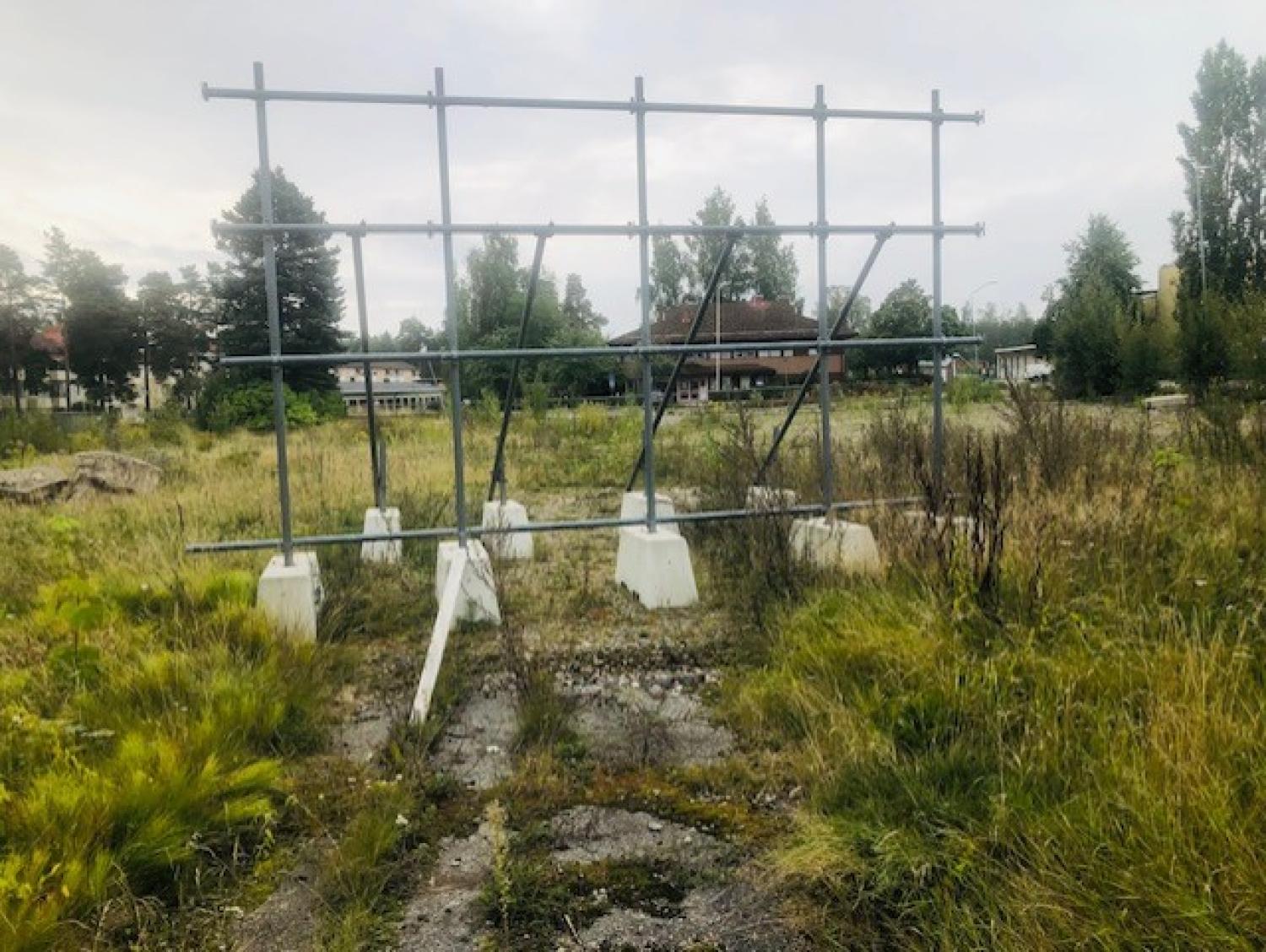 En metallram för en skylt står tom i en övervuxen gräsyta med byggnader och träd i bakgrunden under en molnig himmel.