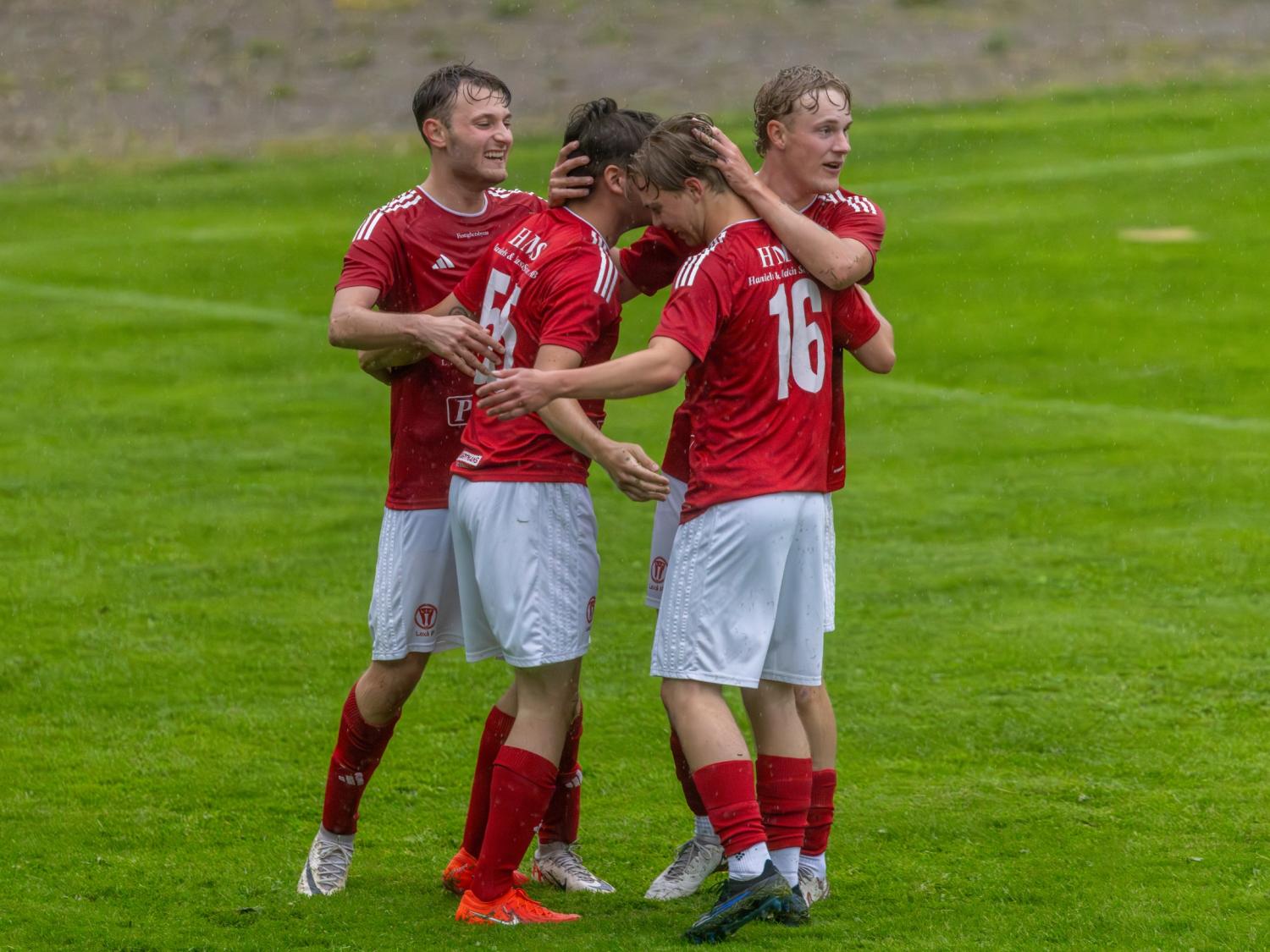 Fyra fotbollsspelare i röda tröjor står på en gräsplan och firar och kramar varandra under en match.
