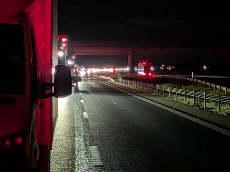 En nattlig motorvägsscen med utryckningsfordon och trafik längre fram, röda ljus syns, vägen är delvis upplyst och en viadukt skymtar i fjärran.