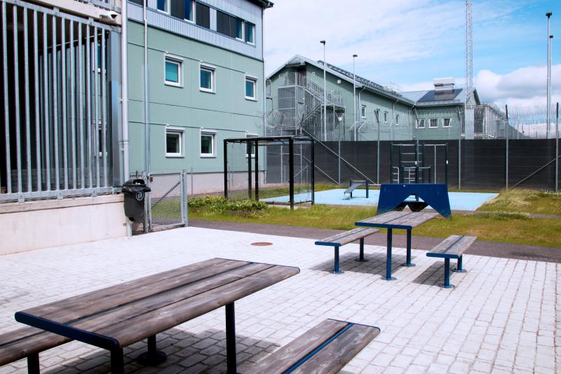Utomhusområdet vid en bevakad anläggning med metallbänkar, träbord, ett pingisbord, en inhägnad gård och omgivande byggnader.