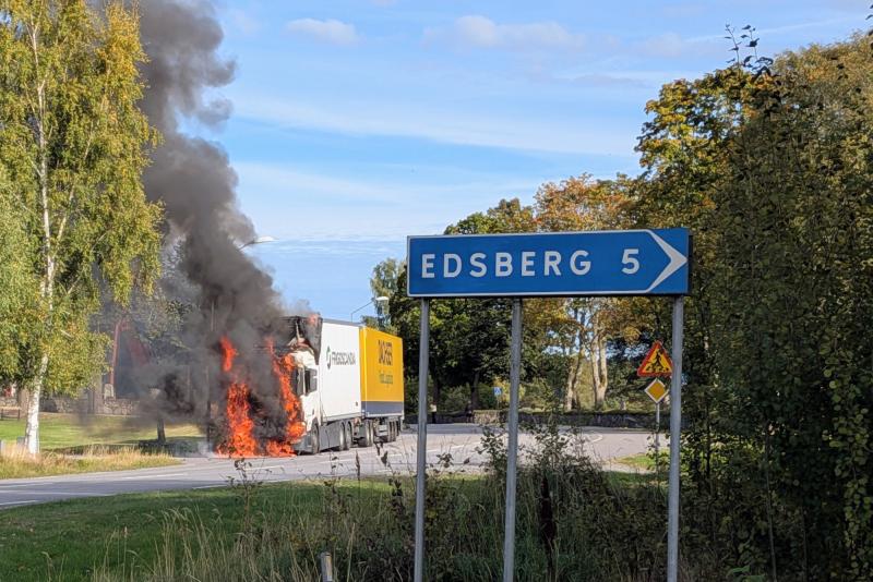 En lastbil som står i brand vid vägkanten sprider svart rök nära en vägskylt som anger att Edsberg ligger 5 kilometer bort.