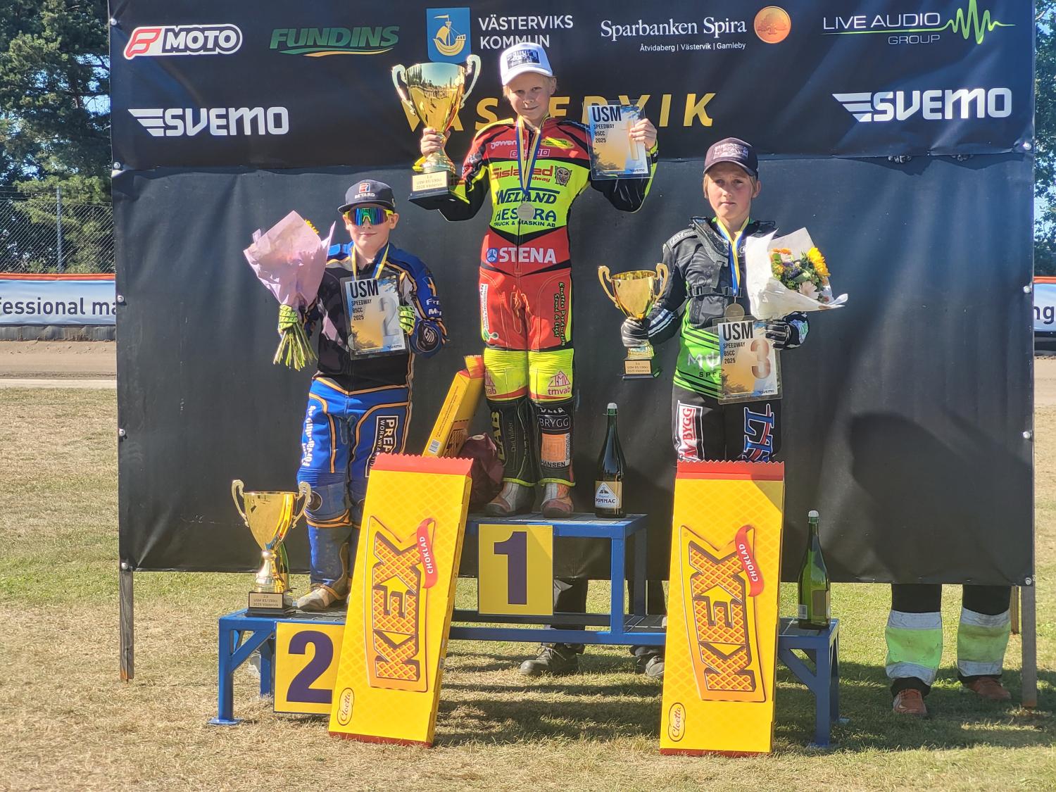 Tre motocrossförare står på ett prispall med pokaler och buketter i händerna, med skyltar för första, andra och tredje plats framför sig.