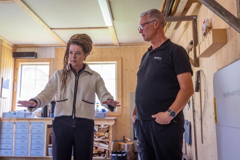 Två personer står i en snickarverkstad; den ena gestikulerar med båda händerna medan den andra tittar uppmärksamt på.