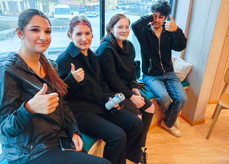 Fyra personer sitter inomhus på en bänk vid ett fönster, tre gör tummen upp och en håller i en burk, alla är mörkklädda.