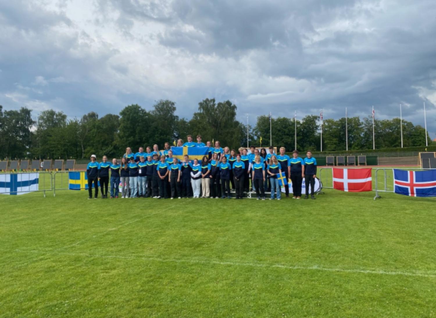 En stor grupp människor står på en gräsbevuxen plan, några håller i en svensk flagga. Flera nationella flaggor inklusive Sveriges, Finlands, Danmarks och Islands flaggor visas framför.
