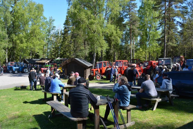 Människor sitter vid picknickbord och står i grupper nära en rad röda och blå traktorer i ett gräsbevuxet, trädbevuxet utomhusområde en solig dag.