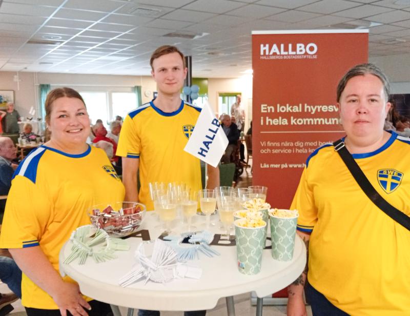 Tre personer i gula svenska fotbollströjor står vid ett bord med drycker, snacks och en 