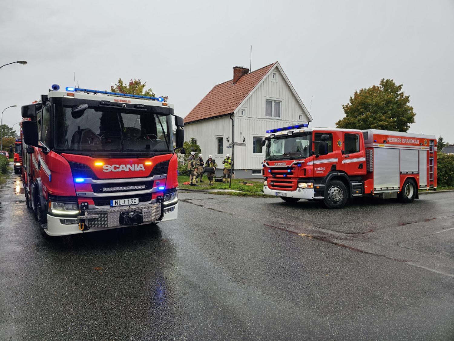 Två brandbilar är parkerade på en våt gata nära ett vitt hus, med flera brandmän som står vid byggnaden under molnig himmel.