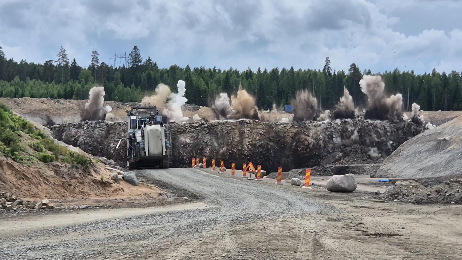 En byggarbetsplats med en grusväg, orange avspärrningar, en stor maskin och en explosion som spränger stenar i bakgrunden, med en skog och ett molnigt himmel.