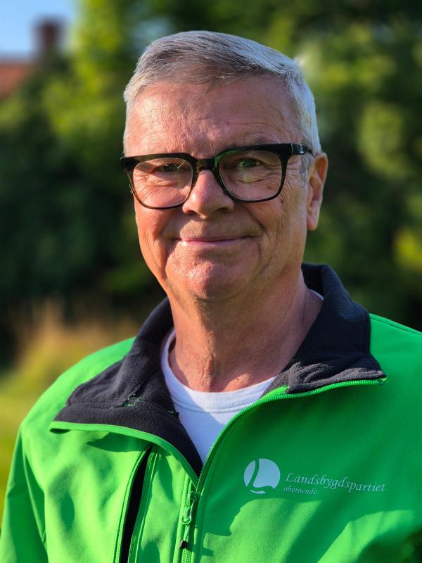 En äldre man med kort grått hår och glasögon bär en ljusgrön jacka med ”Landsbygdspartiet”-logotypen och står utomhus med grönska i bakgrunden.