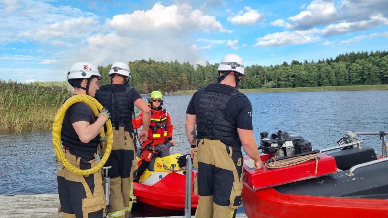Fyra brandmän i skyddsutrustning och hjälmar förbereder en liten röd räddningsbåt vid stranden av en sjö, med träd och vass i bakgrunden, under en delvis molnig himmel.