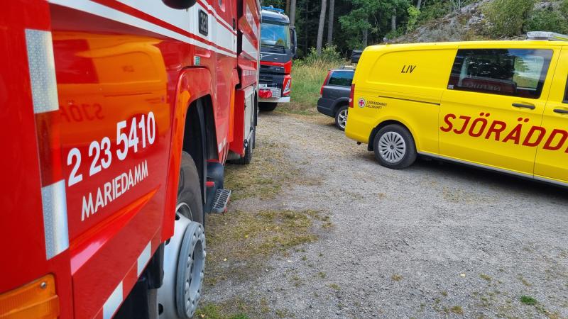 En röd brandbil och ett gult utryckningsfordon med texten ”SJÖRÄDDNING” står parkerade på en grusplan nära träd, vilket tyder på en räddningsinsats.