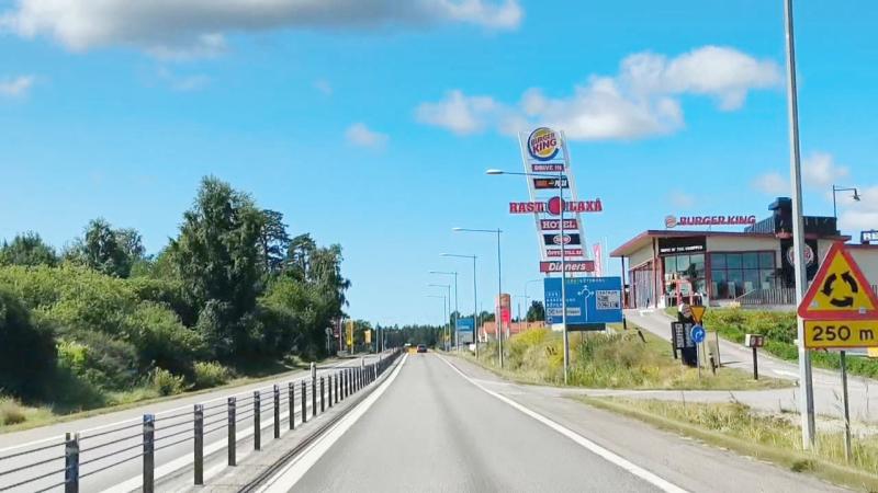 En väg med mitträcke och trafikskyltar, en Burger King-restaurang, skyltar för en bensinstation, träd på vänster sida och en varningsskylt för övergångsställe på höger sida.