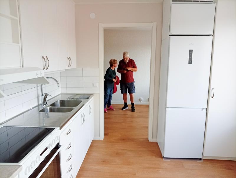 Två personer står och pratar i dörröppningen till ett ljust, modernt kök med vita skåp, elspis och ett stort kylskåp.