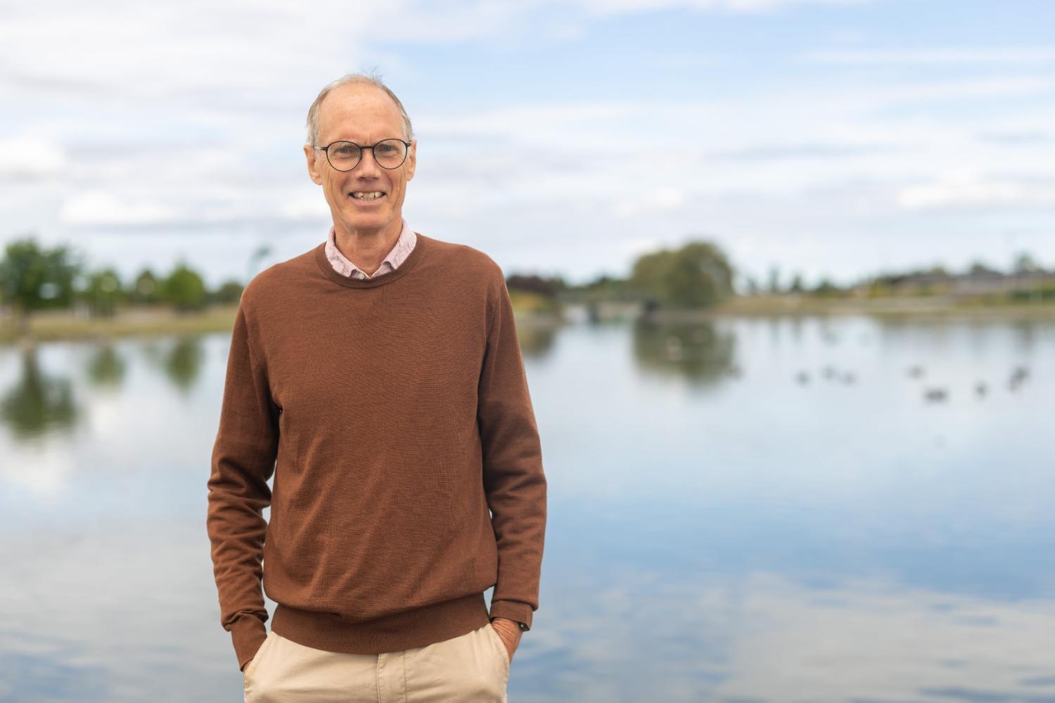 En man med glasögon, en brun tröja och khakifärgade byxor står utomhus nära en stilla sjö, med träd och en molnig himmel i bakgrunden.