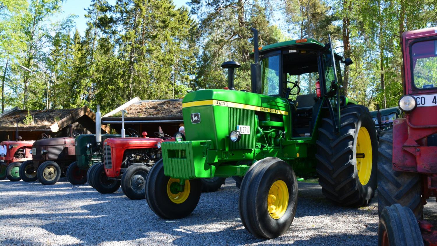 En rad veterantraktorer, däribland en grön John Deere 4250, står parkerade utomhus på en grusplan med träd och träbyggnader i bakgrunden.