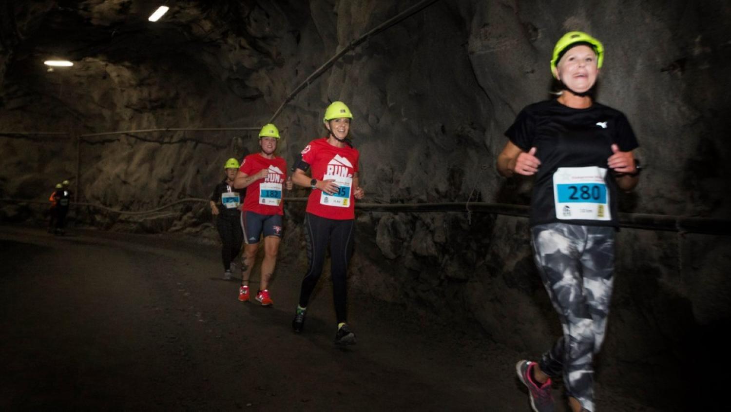 Löpare med pannlampor och numrerade haklappar deltar i ett lopp genom en svagt upplyst tunnel med steniga väggar.