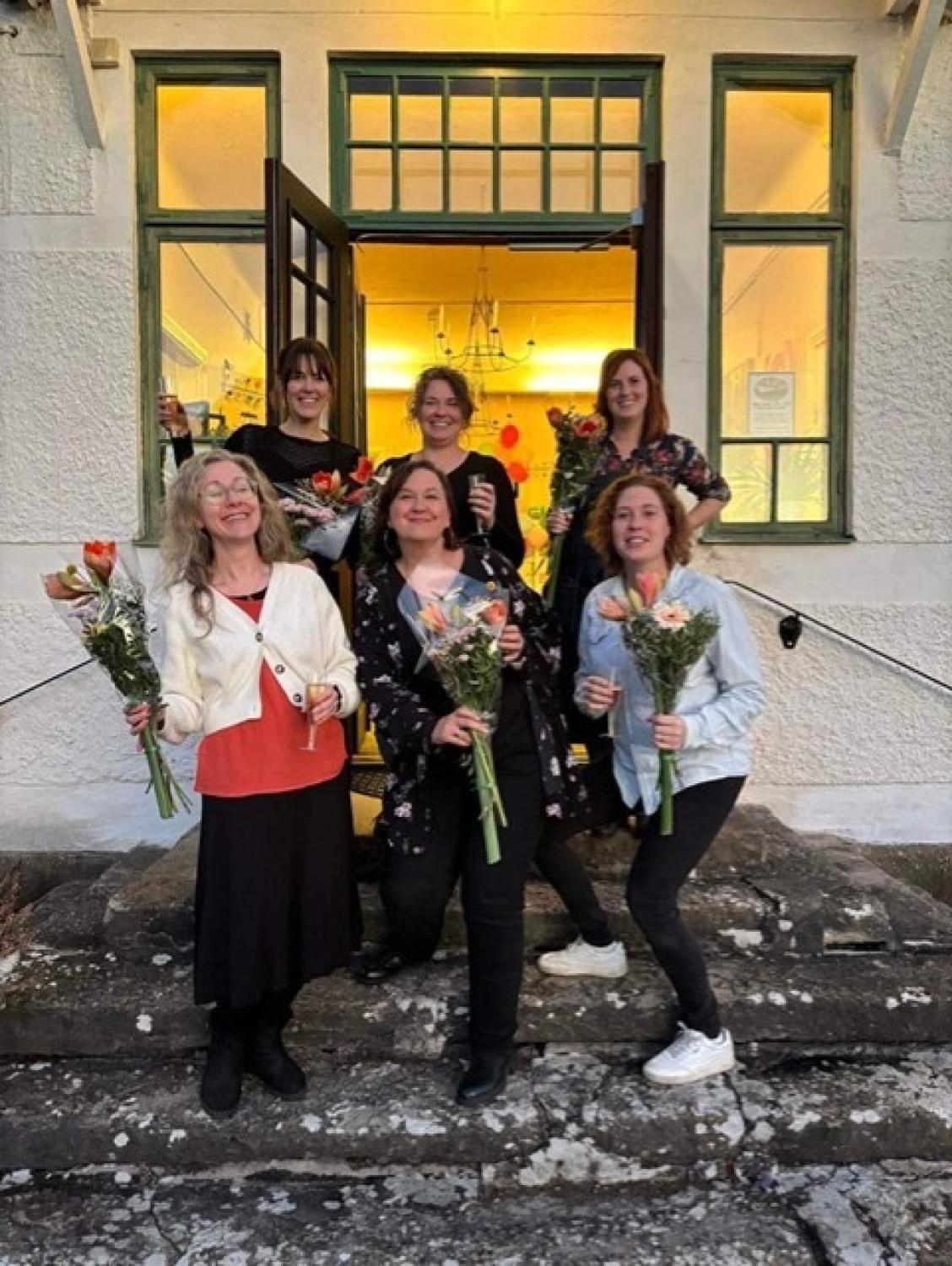 Sex personer står utanför en byggnad, var och en med en bukett blommor och en drink och ler mot kameran.