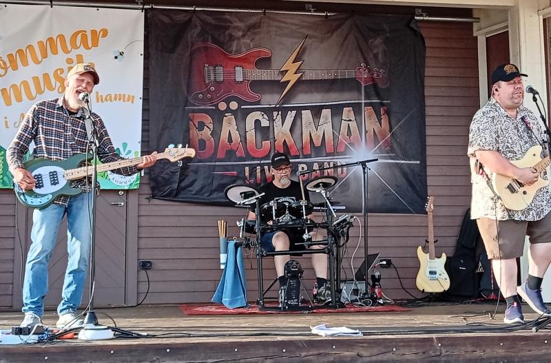 Tre musiker uppträder på en utomhusscen med gitarrer och trummor. På en banderoll bakom dem står det ”Bäckman”. Stämningen verkar avslappnad och ledig.