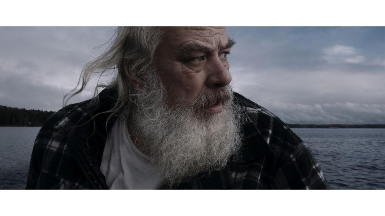 En äldre man med långt vitt skägg och rutig jacka tittar åt sidan när han sitter vid en vattensamling under en molnig himmel.