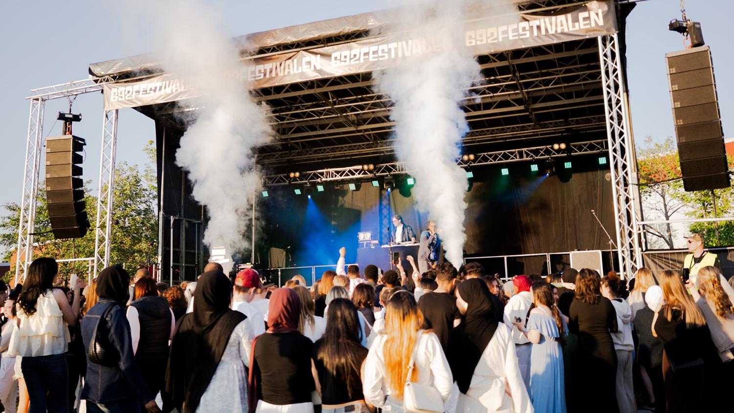 En publik tittar på ett uppträdande på en utomhusscen med rökeffekter, belysning och två personer bakom DJ-utrustning under en skylt med texten 