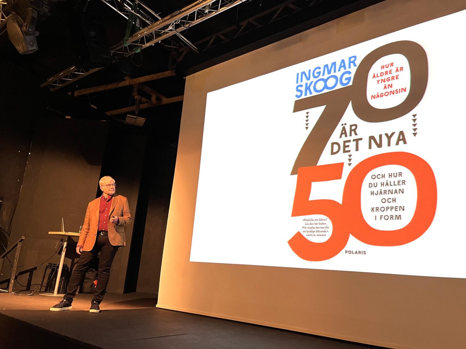 En person står på scenen bredvid ett stort projicerat bokomslag med titeln 