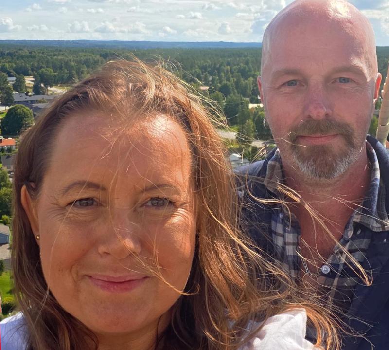 Två personer som står tillsammans utomhus och tittar mot kameran, med grönska och en delvis molnig himmel i bakgrunden.