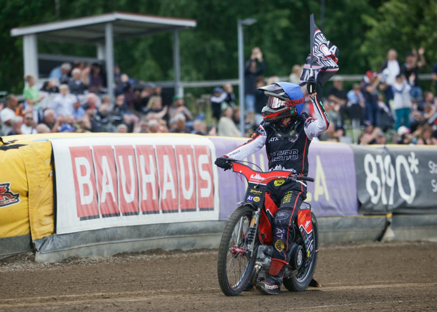 En speedwayförare på en grusbana viftar med en flagga medan han kör förbi publiken, med reklambanderoller synliga i bakgrunden.
