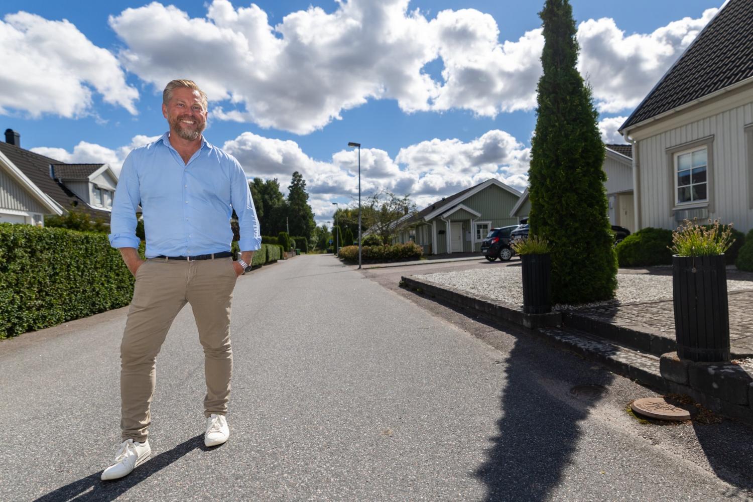 En man i en ljusblå skjorta och beige byxor står och ler på en lugn bostadsgata kantad av hus och grönska under en delvis molnig himmel.