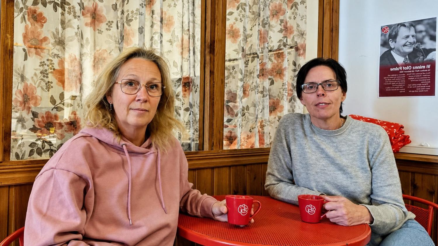 Två kvinnor sitter vid ett rött bord med matchande röda muggar i ett mysigt rum med blommiga gardiner och en affisch på väggen bakom dem.